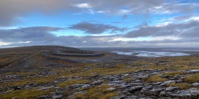 Courtesy Bernice Naughton, Fáilte Ireland, burren mullaghmore loop scenic hiking trail with stunning views of the unique limestone landscape, rolling hills, and diverse flora. Grey clouds covering the blue sky. Dark grey stones and khaki greenery covering the landscape.